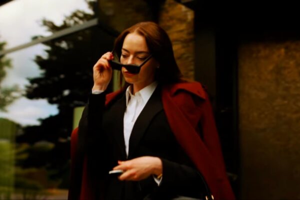 صورة لشخصية أنيقة ترتدي معطفًا أحمر وتنتظر خارج مبنى زجاجي، مع حاملة حقيبة ونظارات في يدها، من فيلم بوغونيا (Bugonia) بطولة Emma Stone.