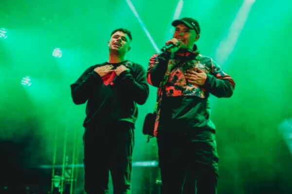 صورة لفرقة Kneecap أثناء أدائهم الموسيقي على المسرح، مع إضاءة خضراء وجمهور متحمس، يظهر Mo Chara وMóglaí Bap وهما يغنيان بحماس.