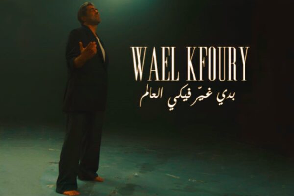 صورة لوائل كفوري (Wael Kfoury) يقف في إضاءة خافتة ببدلة سوداء، مع نص "وائل كفوري" و"بدي غيّر فيكي العالم" مكتوب بالعربية والإنجليزية، من فيديو أغنيته الجديدة.