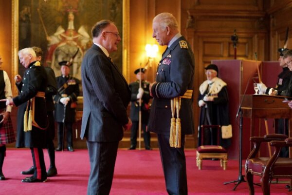 صورة للملك تشارلز الثالث وهو يقدم أوسمة تكريمية لأبطال اسكتلندا في قصر هوليرودهوس، مع حضور أشخاص بزي رسمي تقليدي وحديث في قاعة فاخرة.