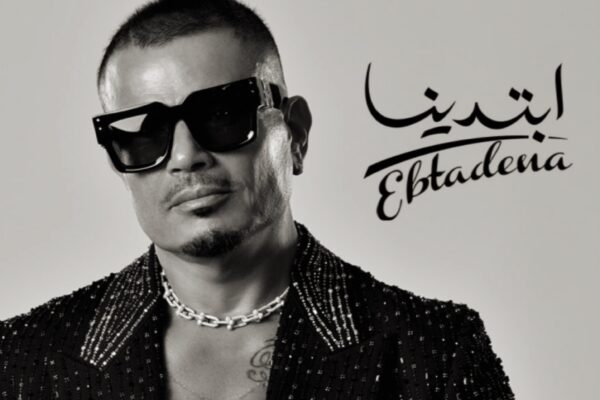 صورة للفنان عمرو دياب يرتدي نظارة شمسية سوداء وسترة مزخرفة، مع كلمة "ابتدينا" مكتوبة بالعربية والإنجليزية إلى جانبه.