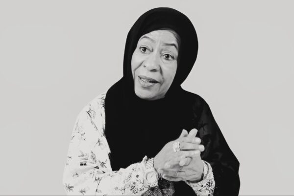 صورة أبيض وأسود للفنانة الإماراتية رزيقة الطارش ترتدي حجابًا أسود وحجابًا مزخرفًا، مع يديها متشابكتين تعكسان أناقتها وإرثها الفني.