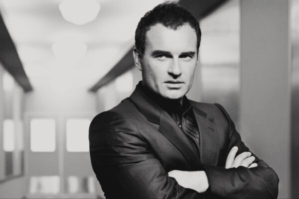 صورة أبيض وأسود للممثل Julian McMahon يرتدي بدلة رسمية ويضع يديه متقاطعتين، مع خلفية داخلية ضبابية.
