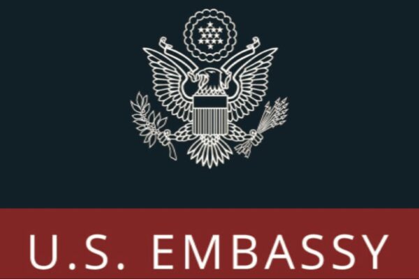 صورة تحتوي على شعار السفارة الأمريكية الرسمي، مع نسر أبيض ودرع على خلفية داكنة، مصحوبًا بلافتة حمراء تحمل النص "U.S. EMBASSY" باللون الأبيض.