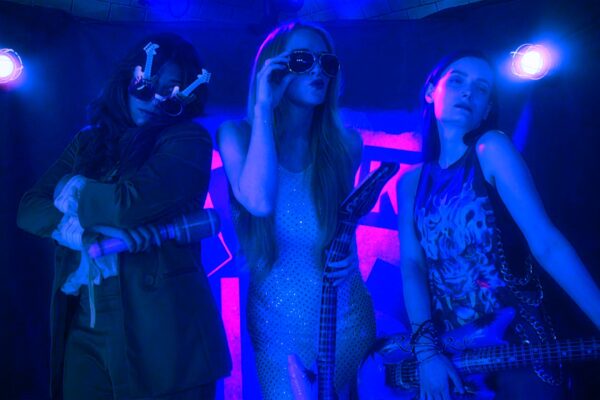 ثلاث موسيقيين يعزفون على المسرح تحت أضواء زرقاء وبنفسجية، واحد يعزف على الجيتار، والثاني يمسك الميكروفون، والثالث مع جيتار، جميعهم بملابس أنيقة. Three musicians performing on stage under blue and purple lighting, with one playing a guitar, another holding a microphone, and the third with a guitar, all dressed in stylish outfits.