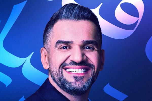 صورة للفنان حسين الجسمي يبتسم أمام خلفية زرقاء تحتوي على كلمات عربية بألوان متدرجة.