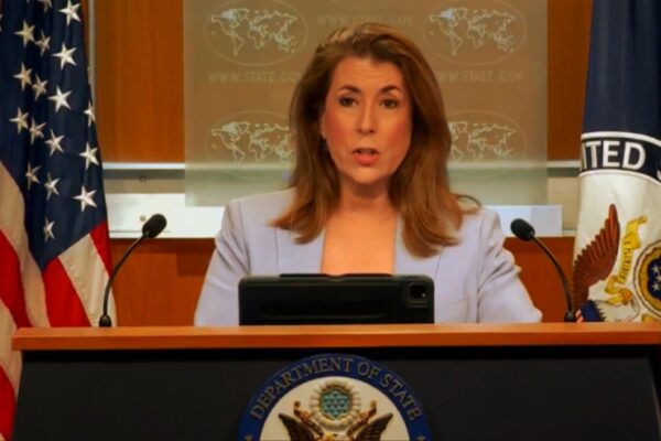 صورة تُظهر متحدثة باسم وزارة الخارجية الأمريكية تقف خلف منصة تحمل شعار "Department of State"، مع أعلام الولايات المتحدة ووزارة الخارجية في الخلفية، أثناء إلقاء تصريح رسمي.