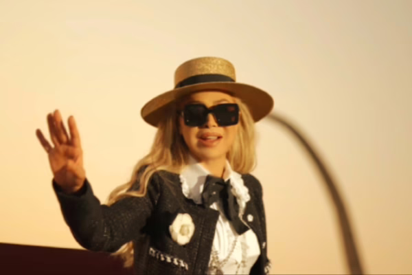 صورة لنوال الزغبي ترتدي قبعة قش ونظارات شمسية سوداء، وهي تلوح بيدها بابتسامة، مرتدية جاكيت أسود مزين بتفاصيل بيضاء، مع خلفية دافئة تعكس ضوء الغروب.