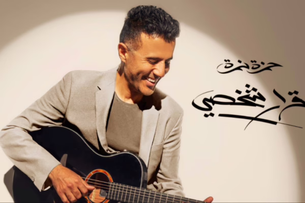 صورة لحمزة نمرة وهو يعزف على الجيتار بابتسامة مشرقة، مرتديًا بدلة رمادية، مع خلفية بيضاء تحمل توقيعه بخط عربي أنيق.