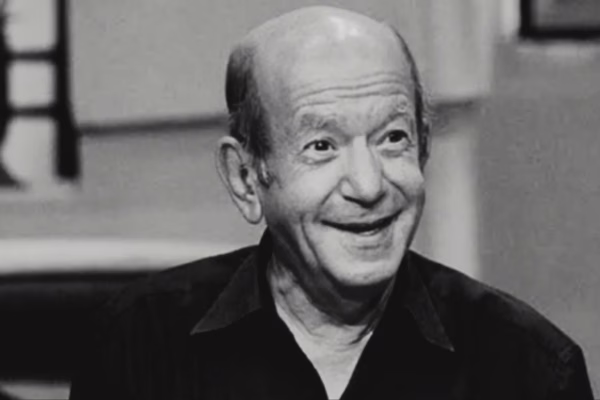 صورة أبيض وأسود للفنان المصري لطفي لبيب، يظهر وهو يبتسم بثقة، مرتديًا قميصًا داكنًا، مع خلفية تحتوي على أثاث بسيط وإضاءة خافتة.