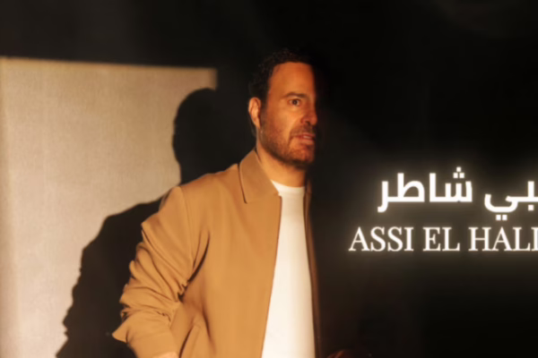 صورة عاصي الحلاني في جلسة تصوير مميزة لأغنية قلبي شاطر، يرتدي جاكيت بني مع خلفية مظلمة ونص مضيء يعكس عنوان الأغنية.