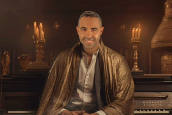 صورة للمطرب محمد عدوية يجلس أمام بيانو في بيئة دافئة مضاءة بشموع، مرتديًا عباءة ذهبية أنيقة، مع خلفية مزينة بزخارف شرقية تبرز أصالته الفنية.