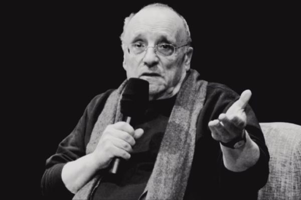 صورة بالأبيض والأسود لـ Marcel Łoziński، مخرج وثائقي بولندي، وهو يتحدث إلى ميكروفون بيده مرفوعة، مرتديًا نظارات ووشاح.