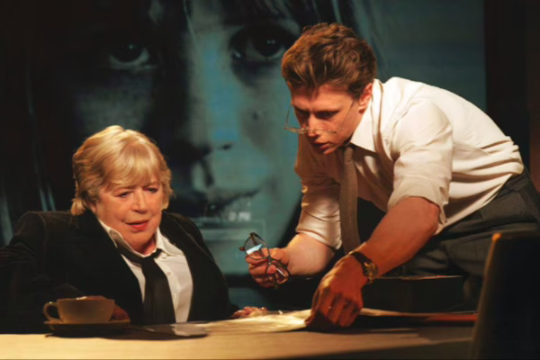 صورة تجمع بين امرأتين تجلسان خلف مكتب وشاب يقدم نظارات، مع خلفية تحمل صورة وجه ضخمة، تعكس أجواء الفيلم السينمائي المؤثر لماريان فيثفول.