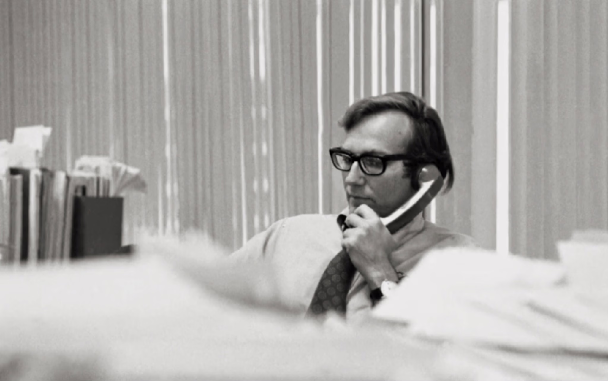 لقطة أرشيفية بالأبيض والأسود من فيلم Cover-Up تظهر الصحفي الشاب سيمور هيرش جالساً في مكتبه، يرتدي نظارته الطبية ويتحدث عبر الهاتف بتركيز شديد وسط أكوام من الأوراق، مما يعكس أجواء العمل الصحفي الاستقصائي في تلك الحقبة.