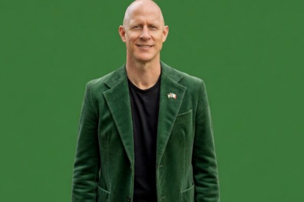 بورتريه رسمي لبرايان ماكغينيس (Brian McGinnis)، مرشح مجلس الشيوخ والمحارب القديم، يبتسم أمام خلفية خضراء، يرتدي سترة مخملية خضراء ودبوس العلم الفلسطيني على ياقته.