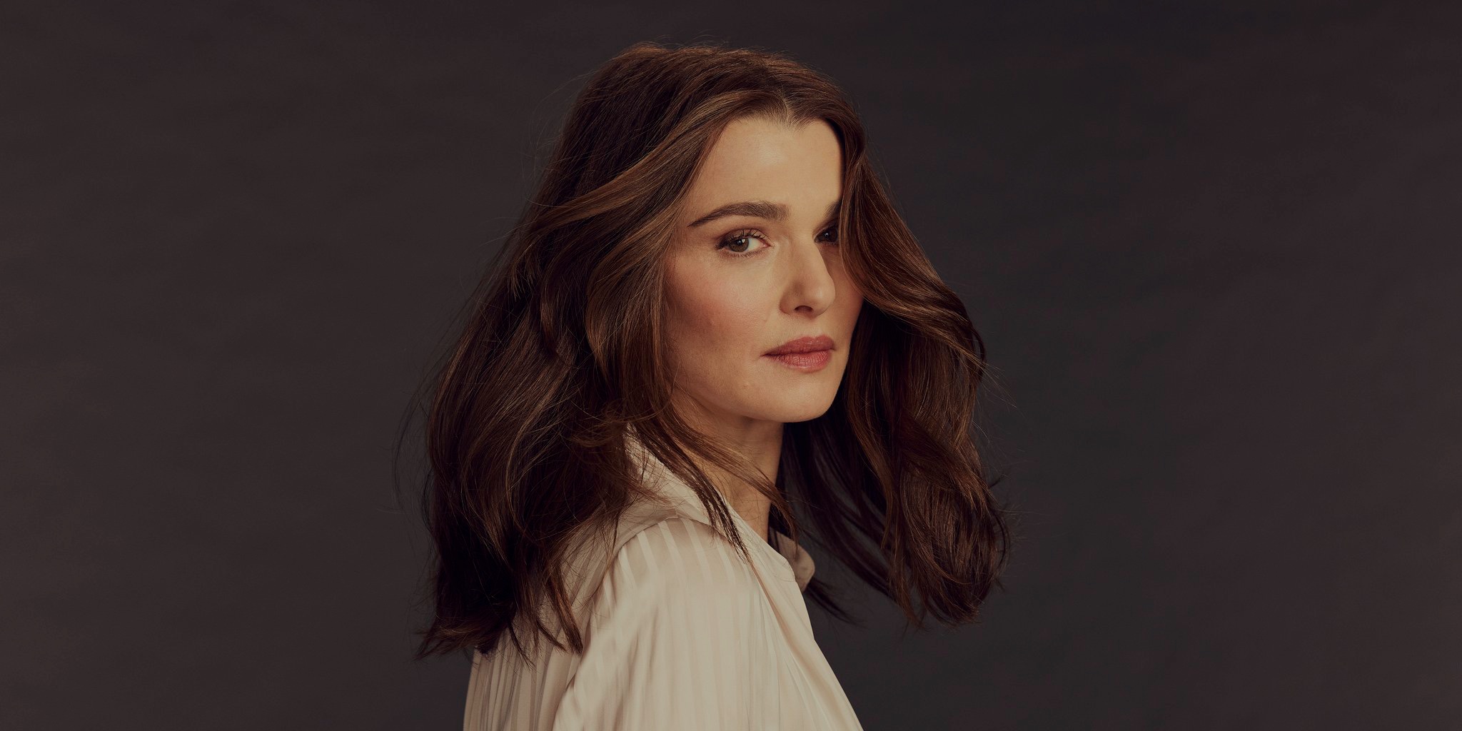 بورتريه جانبي جزئي للممثلة ريتشل وايز (Rachel Weisz) بشعر بني مموج ينسدل على كتفيها، ترتدي قميصًا حريريًا أبيض مطويًا وتنظر مباشرة إلى الكاميرا أمام خلفية رمادية داكنة ناعمة.