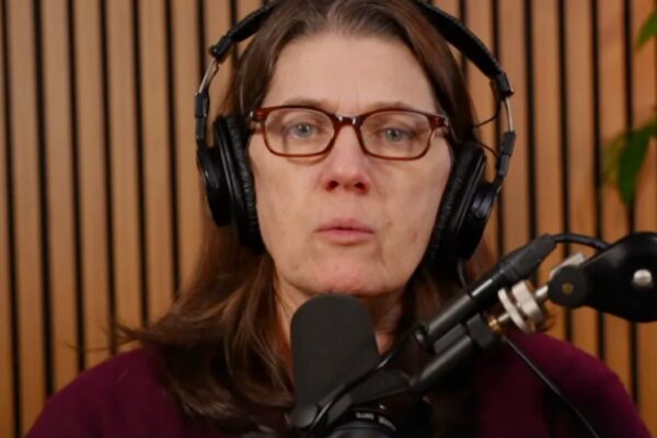 لقطة مقربة لماري ترامب (Mary Trump) ترتدي نظارات طبية وسماعات رأس كبيرة، وتتحدث مباشرة في ميكروفون احترافي (بث مباشر) أمام خلفية من الخشب الطولي.