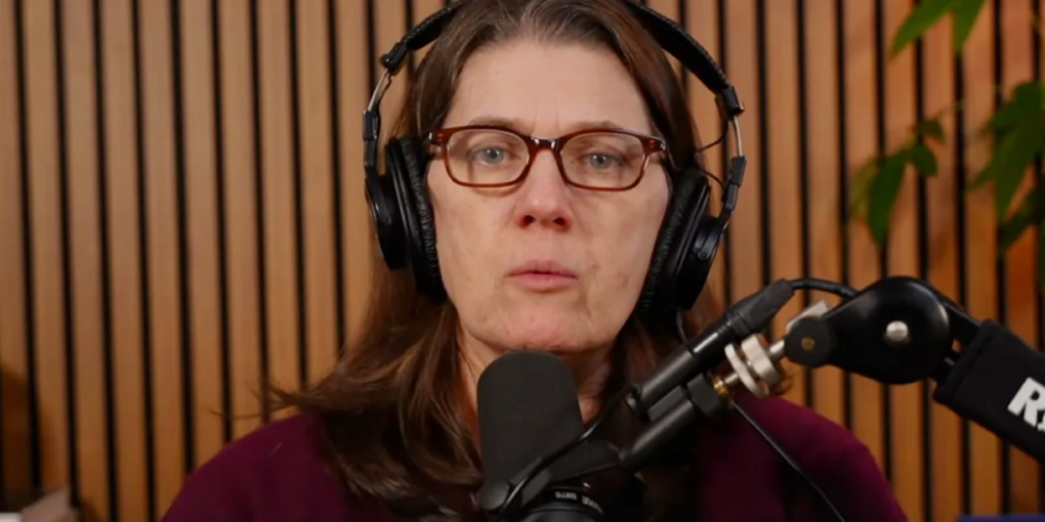 لقطة مقربة لماري ترامب (Mary Trump) ترتدي نظارات طبية وسماعات رأس كبيرة، وتتحدث مباشرة في ميكروفون احترافي (بث مباشر) أمام خلفية من الخشب الطولي.