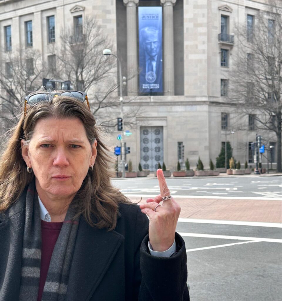 ماري ترامب (Mary Trump) تقف في الشارع أمام مبنى وزارة العدل الأمريكية، وعلى واجهة المبنى لافتة ضخمة تحمل صورة دونالد ترامب؛ ماري تظهر بملامح وجه حادة ومتجهمة وهي تقوم بحركة احتجاجية هجومية بإصبعها الأوسط تجاه الكاميرا/المبنى.