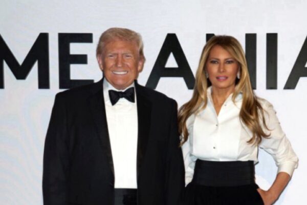 دونالد وميلانيا ترامب يقفان معاً أمام لوحة بيضاء ضخمة تحمل اسمها، للترويج لإصدار فيلم ميلانيا ترامب الوثائقي، حيث يرتدي دونالد بدلة توكسيدو كلاسيكية وترتدي ميلانيا قميصاً أبيض أنيقاً.