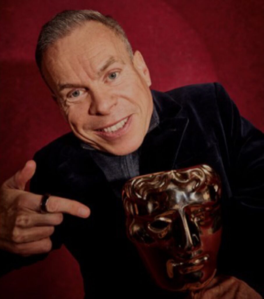 لقطة قريبة للنجم البريطاني وارويك ديفيس وهو يبتسم للكاميرا بحماس ويمسك بتمثال جائزة البافتا الذهبية اللامعة في يده اليسرى، بينما يشير إليها بسبابته اليمنى. يرتدي سترة مخملية زرقاء داكنة (بليزر) فوق قميص أزرق فاتح، وتقف خلفه خلفية منسوجة باللون الأحمر الداكن تشبه السجادة الحمراء أو ستائر المسرح.