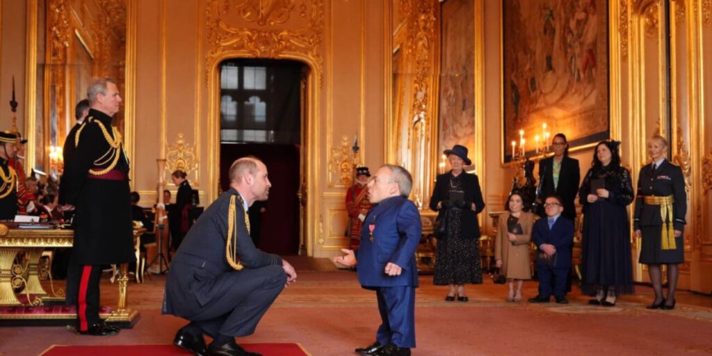 لقطة مؤثرة تجسد الاحترام والتقدير حيث ينحني الأمير وليام ليتحدث بمستوى عيني النجم وارويك ديفيس أثناء مراسم تقليده وسام OBE، وتظهر في الخلفية القاعة المذهبة الفاخرة بالقصر مع تواجد أفراد عائلته ومسؤولي المراسم.