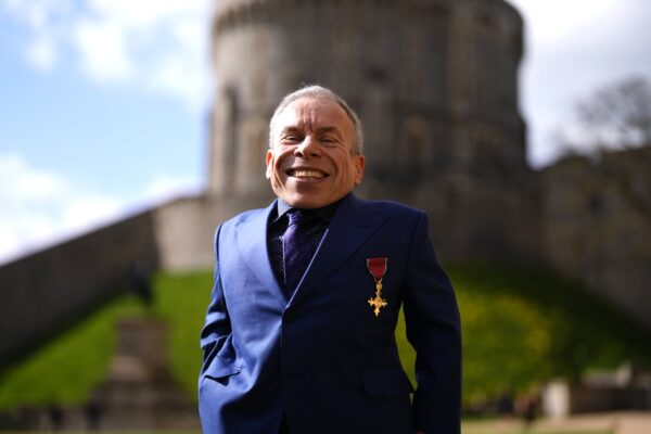 الممثل وارويك ديفيس يبتسم مرتدياً حلة زرقاء ووسام OBE على صدره أمام قصر ويندسور التاريخي في يوم منحه التكريم الملكي.