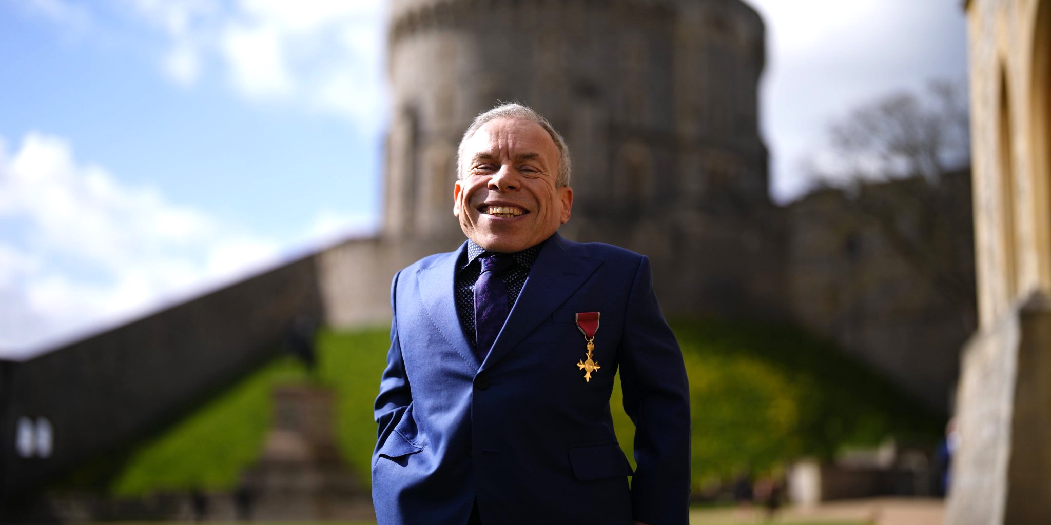 الممثل وارويك ديفيس يبتسم مرتدياً حلة زرقاء ووسام OBE على صدره أمام قصر ويندسور التاريخي في يوم منحه التكريم الملكي.
