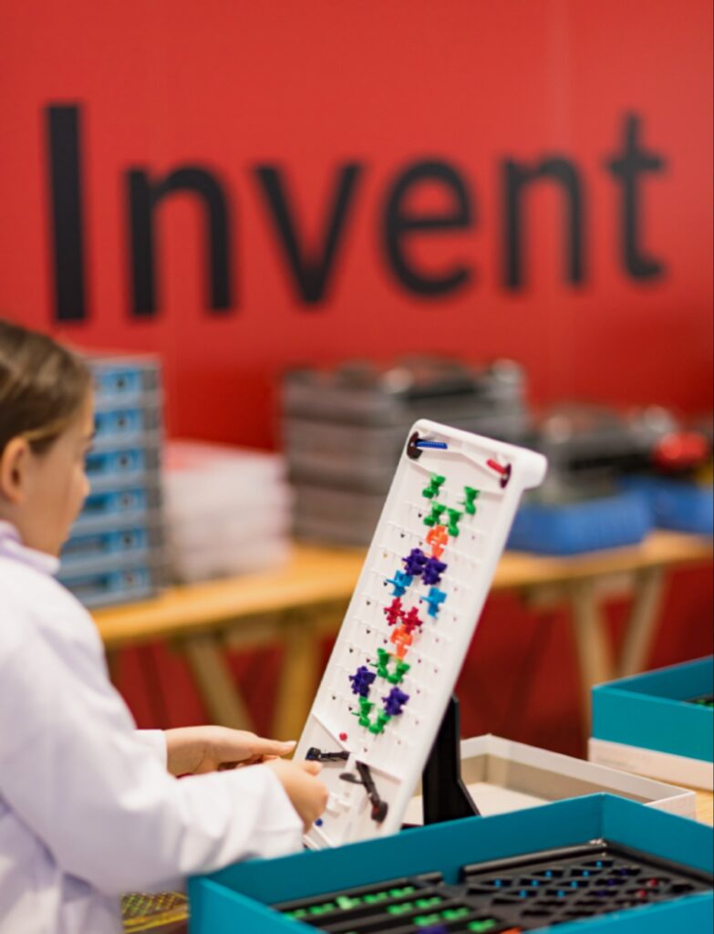 طفلة في "مختبر الشباب" ترتدي معطفاً أبيض وتعمل على لوحة تركيب تعليمية ملونة أمام لافتة "Invent" في معرض جنيف الدولي للاختراعات.