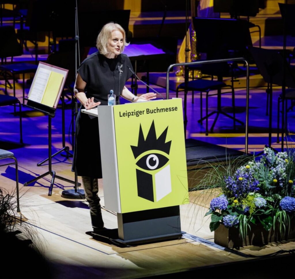 Astrid Böhmisch، مديرة معرض لايبزيغ للكتاب، تلقي كلمتها من على المنصة الصفراء إيذاناً بـ انطلاق معرض لايبزيغ للكتاب اليوم في قاعة Gewandhaus المزدحمة.