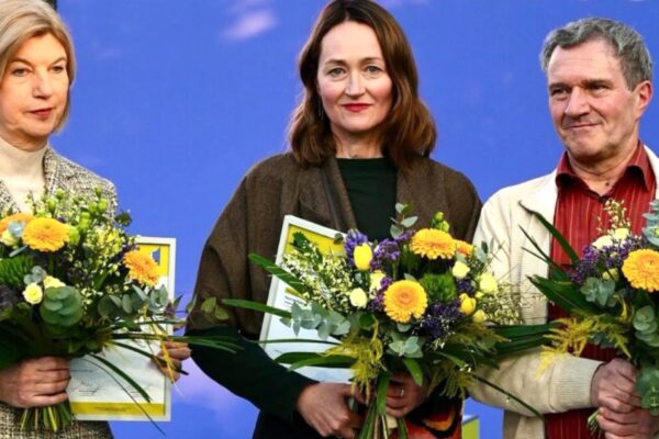 مجموعة صور مجمعة تظهر ثلاثة فائزين مختلفين في لقطات مقربة، كل واحد منهم يحمل باقة زهور كبيرة ووثيقة أو نسخة من كتابه. اللقطة الكبرى (يسار) هي لـ Katerina Poladjan، واللقطتان الأصغر (يمين) هما لـ Marie-Janine Calic و Manfred Gmeiner. الخلفية عبارة عن شاشة زرقاء.