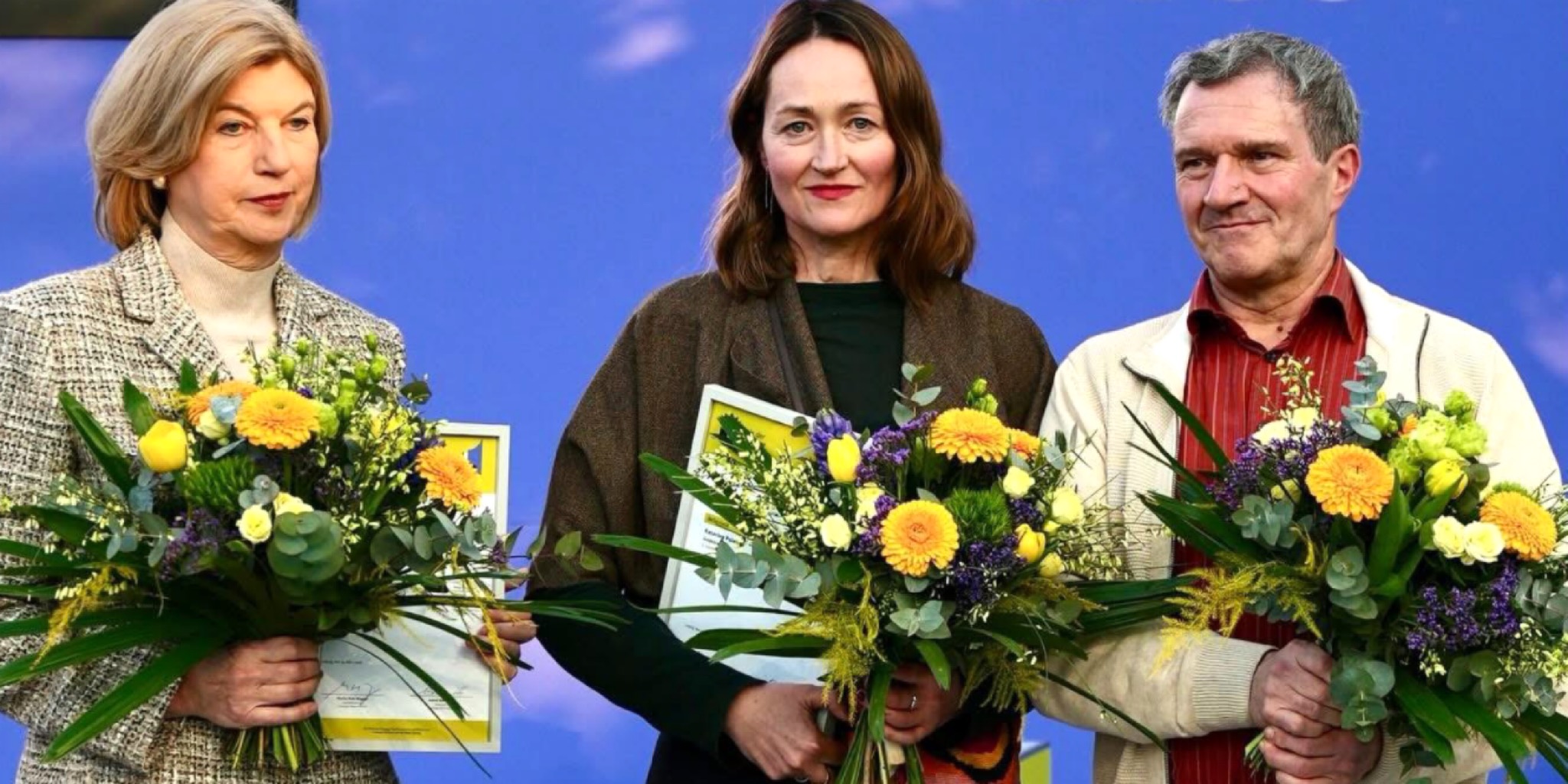 مجموعة صور مجمعة تظهر ثلاثة فائزين مختلفين في لقطات مقربة، كل واحد منهم يحمل باقة زهور كبيرة ووثيقة أو نسخة من كتابه. اللقطة الكبرى (يسار) هي لـ Katerina Poladjan، واللقطتان الأصغر (يمين) هما لـ Marie-Janine Calic و Manfred Gmeiner. الخلفية عبارة عن شاشة زرقاء.