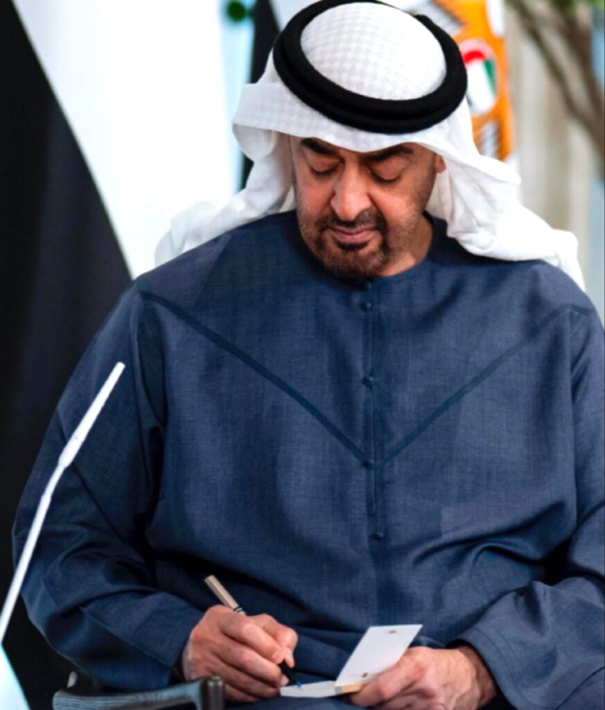 صورة شخصية لصاحب السمو الشيخ محمد بن زايد آل نهيان، يرتدي الكندورة الزرقاء والغترة البيضاء، وهو يركز في الكتابة بيده على بطاقة صغيرة.