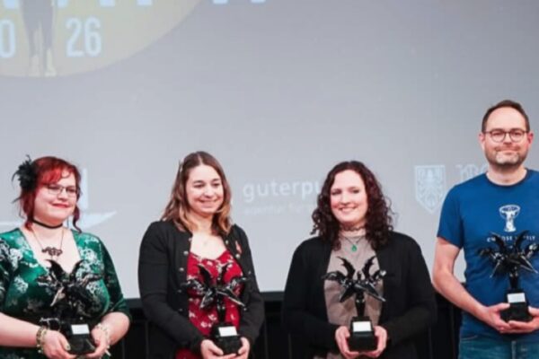 صورة تجمع أربعة فائزين (ثلاث نساء ورجل) يقفون على المسرح ويحملون كؤوس التنانين المجنحة المتميزة، أمام شاشة عرض كبيرة تحمل الرقم '20 26' وشعار جوائز الفانتازيا (Phantastikpreis).