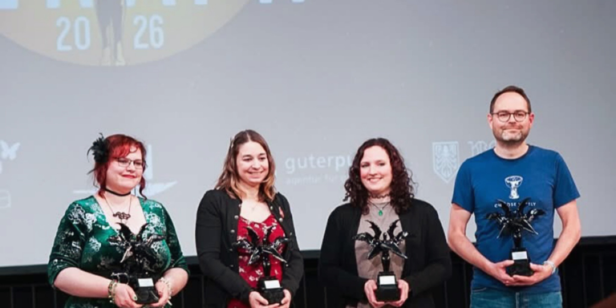 صورة تجمع أربعة فائزين (ثلاث نساء ورجل) يقفون على المسرح ويحملون كؤوس التنانين المجنحة المتميزة، أمام شاشة عرض كبيرة تحمل الرقم '20 26' وشعار جوائز الفانتازيا (Phantastikpreis).