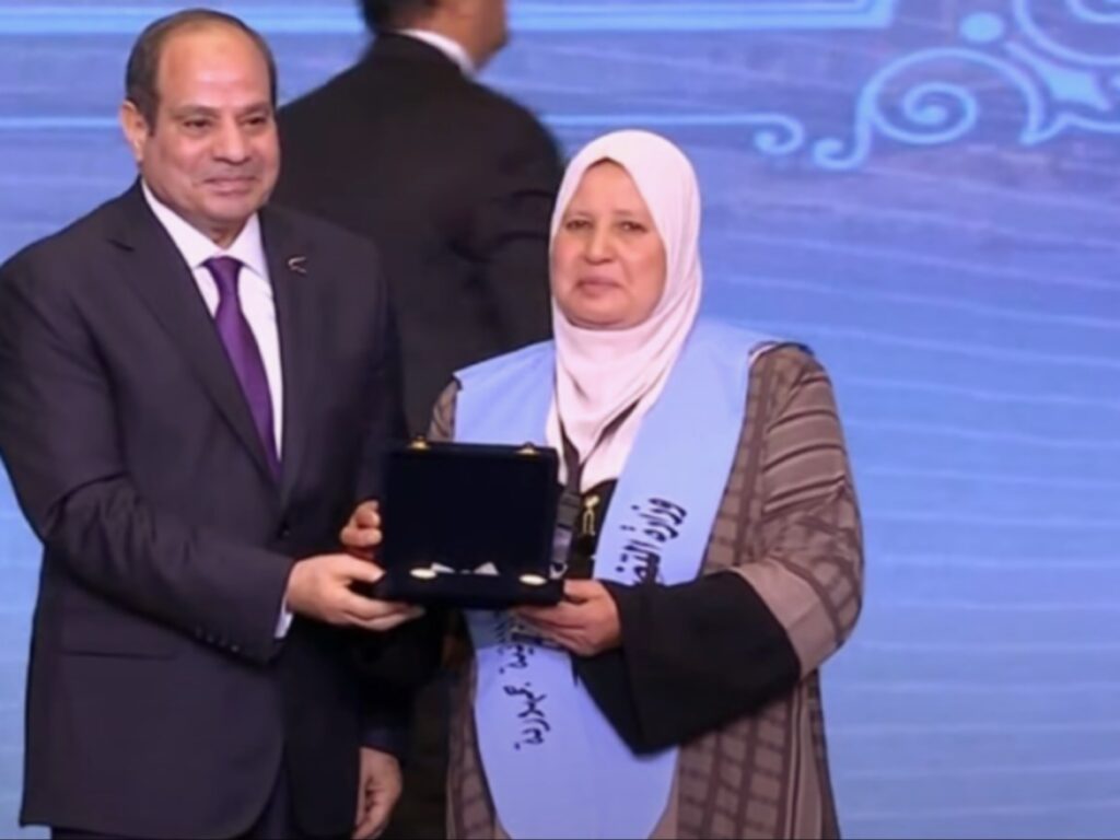 صورة تظهر فخامة الرئيس عبد الفتاح السيسي يقدم جائزة في علبة مفتوحة لسيدة ترتدي الحجاب ووشاحاً أزرق عليه نص عربي، خلال تكريم رسمي.