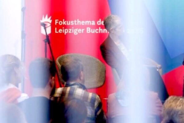 لقطة توثق جلسة نقاشية حية في معرض لايبزيغ للكتاب 2026، تظهر فيها الخلفية الحمراء التي تحمل عبارة "Fokusthema" (الموضوع الرئيسي للمعرض)، مع ظهور متحدثين على المنصة وجمهور يتابع الفعاليات في القاعة رقم 4.