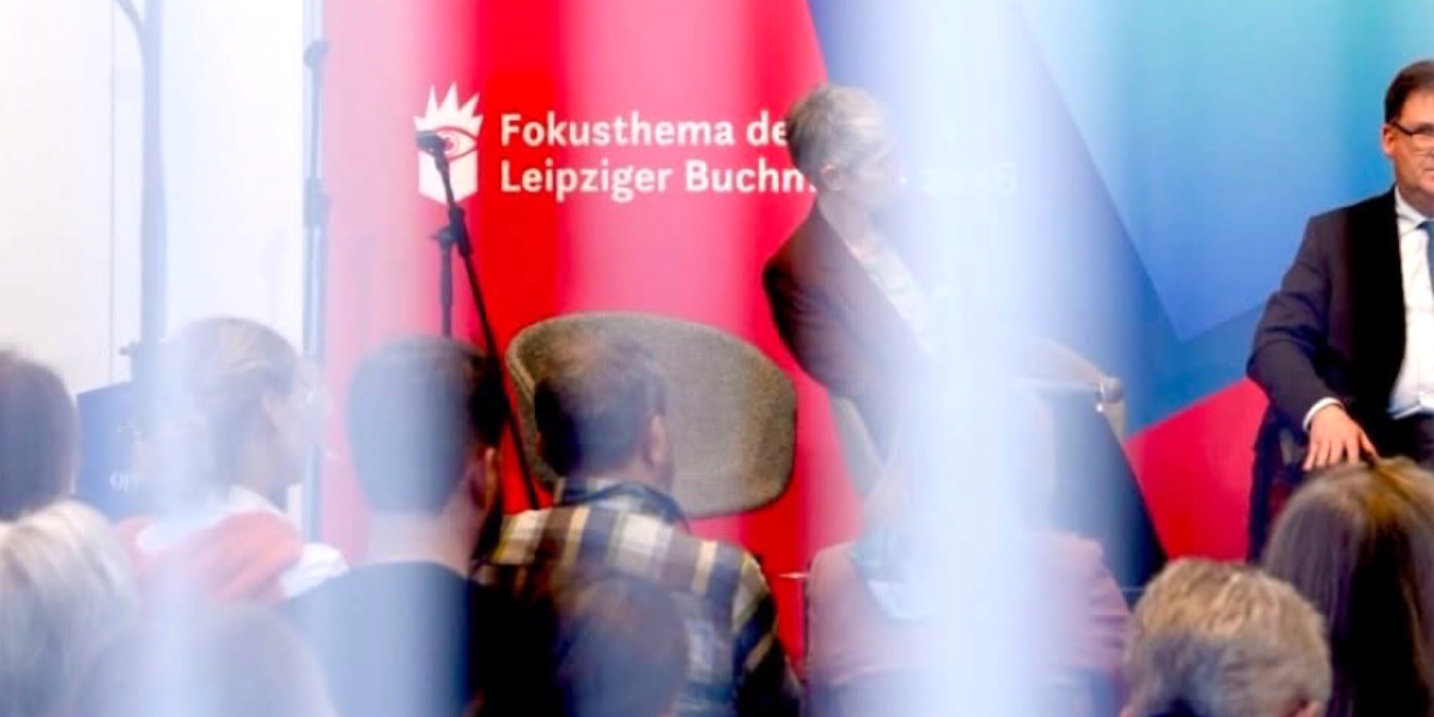 لقطة توثق جلسة نقاشية حية في معرض لايبزيغ للكتاب 2026، تظهر فيها الخلفية الحمراء التي تحمل عبارة "Fokusthema" (الموضوع الرئيسي للمعرض)، مع ظهور متحدثين على المنصة وجمهور يتابع الفعاليات في القاعة رقم 4.