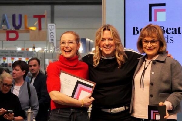 ثلاث سيدات يبتسمن ببهجة على منصة جوائز "BookBeat" في معرض لايبزيغ للكتاب 2026؛ إحداهن تمسك بشهادة تكريم والأخرى تحمل جائزة مكعبة سوداء، وخلفهن شاشة رقمية كبيرة تعرض شعار "BookBeat Awards" وسط أجواء احتفالية وجمهور يتابع الحدث.