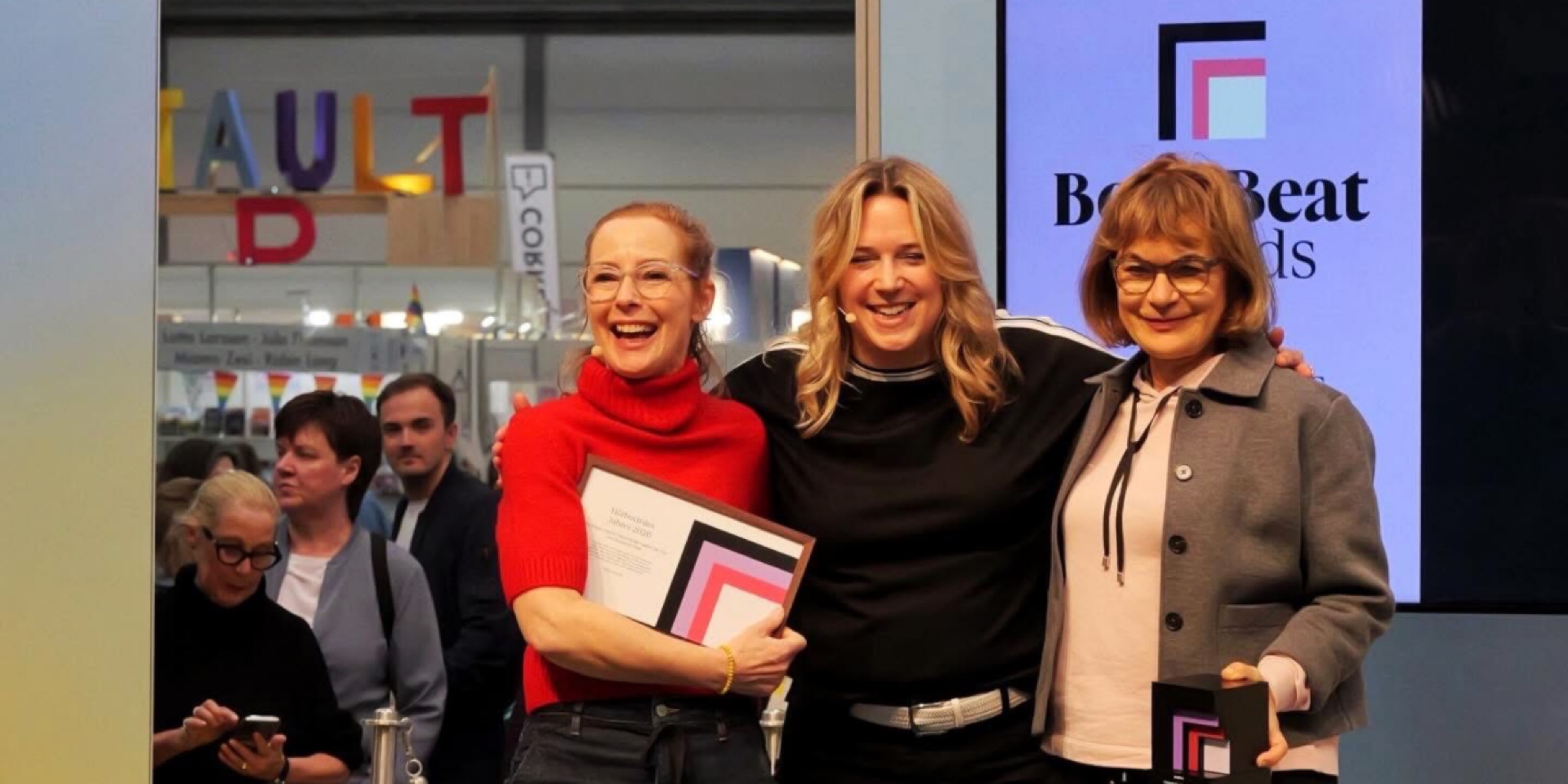 ثلاث سيدات يبتسمن ببهجة على منصة جوائز "BookBeat" في معرض لايبزيغ للكتاب 2026؛ إحداهن تمسك بشهادة تكريم والأخرى تحمل جائزة مكعبة سوداء، وخلفهن شاشة رقمية كبيرة تعرض شعار "BookBeat Awards" وسط أجواء احتفالية وجمهور يتابع الحدث.