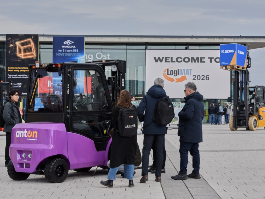 لقطة خارجية تظهر رافعة شوكية أرجوانية من ماركة "anton" في المقدمة، ومجموعة من الناس يقفون أمام مبنى معارض حديث. لافتة كبيرة على المبنى تحمل عبارة 'WELCOME TO LogiMAT 2026' مع شعار المعرض.