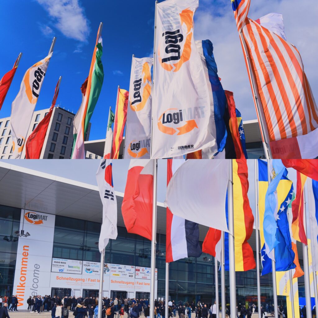 أعلام دولية ترفرف أمام مبنى كبير في يوم مشمس مع حشد من الناس وبوابة مكتوب عليها 'Willkommen'.