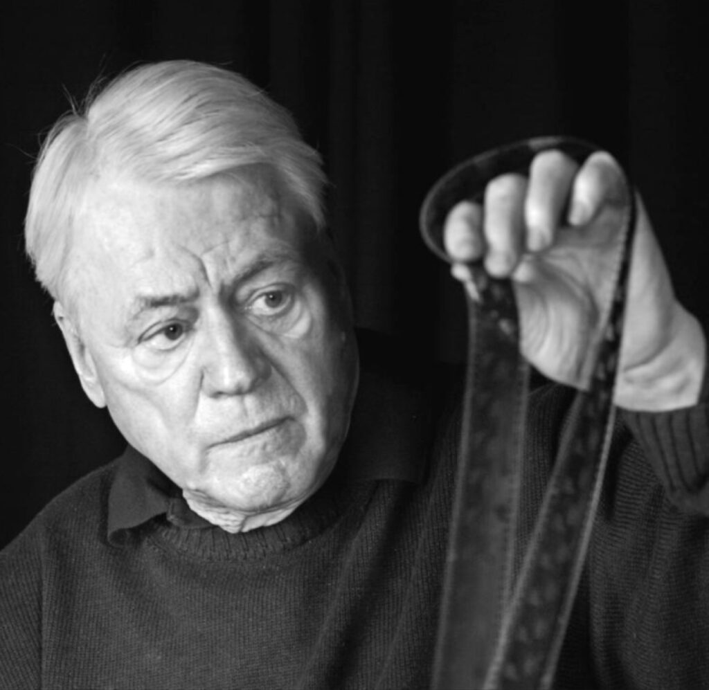 لقطة مقربة بالأبيض والأسود يظهر فيها ألكسندر كلوجه وهو يتفحص شريطاً سينمائياً بتركيز شديد، مما يعكس أسلوبه التجريبي في السرد البصري.