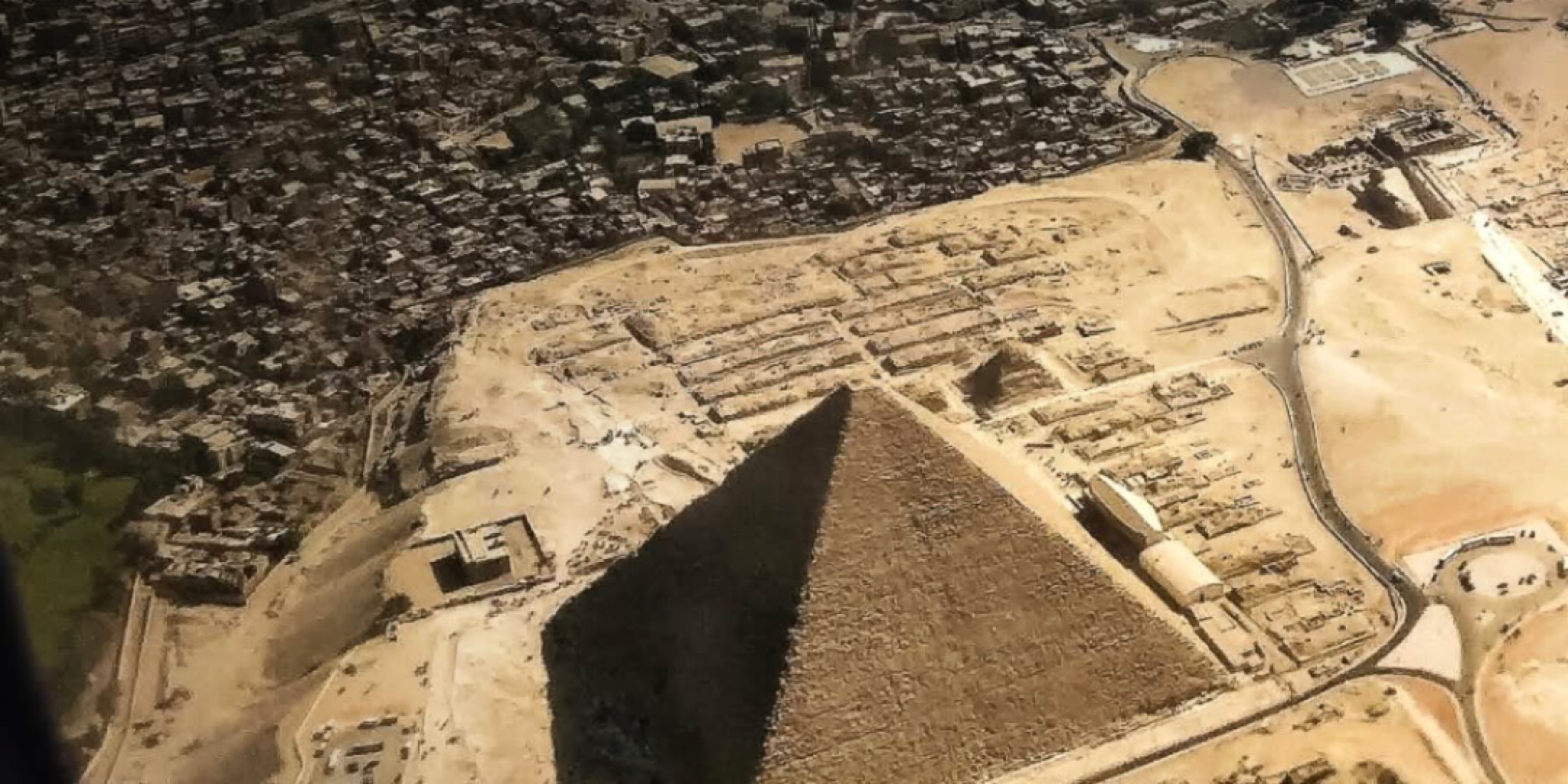 لقطة جوية من نافذة طائرة تظهر هرم خوفو الأكبر ومنطقة الأهرامات في الجيزة مع الكتلة السكنية المحيطة بالقاهرة تحت أشعة الشمس.
