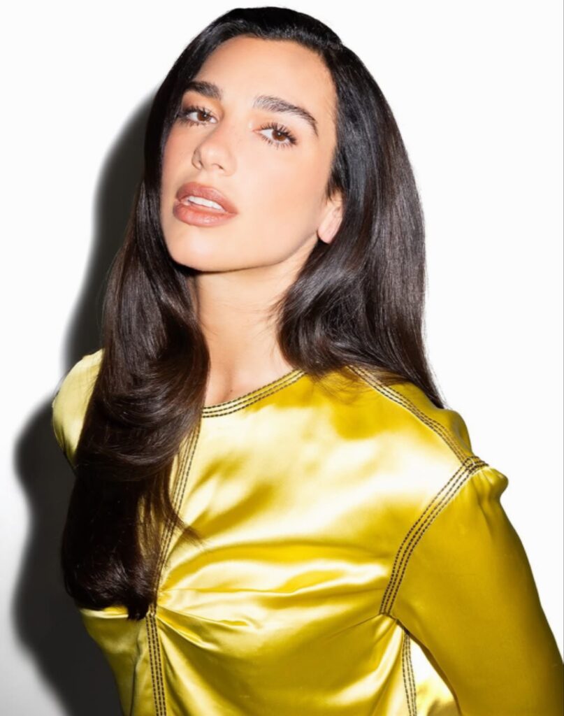 بورتيه شخصي للمغنية العالمية دوا ليبا (Dua Lipa) وهي تنظر للأعلى قليلاً بملامح تأملية هادئة، ترتدي قميصاً من الستان الأصفر اللامع، مع شعر داكن طويل وخلفية بيضاء ناصعة؛ وهي الصورة الرسمية المعتمدة لإعلان مشاركتها في مهرجان London Literature Festival.