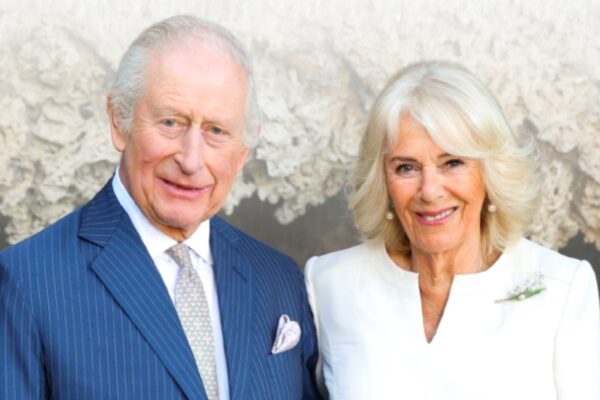 صورة لجلالة الملك تشارلز الثالث في حلة زرقاء مخططة ومنديل جيب وردي، وجلالة الملكة كاميلا في فستان أبيض وعقد لؤلؤ ودبوس زهرة، يبتسمان أمام خلفية طبيعية صخرية.