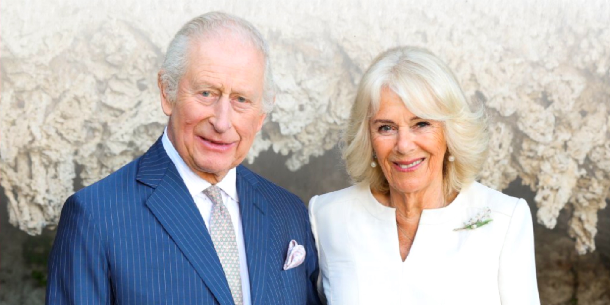صورة لجلالة الملك تشارلز الثالث في حلة زرقاء مخططة ومنديل جيب وردي، وجلالة الملكة كاميلا في فستان أبيض وعقد لؤلؤ ودبوس زهرة، يبتسمان أمام خلفية طبيعية صخرية.