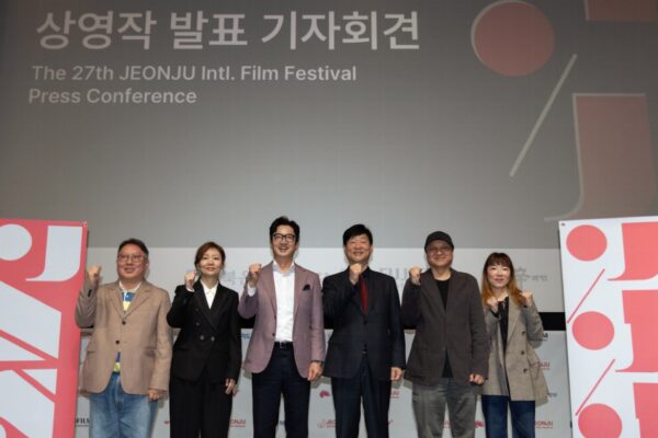لقطة واسعة من الأمام تظهر ستة أشخاص واقفين على خشبة مسرح بملابس رسمية وبذلات، يرفعون قبضات أيديهم اليسرى عالياً في لفتة تأييد جماعية، أمام شاشة ضخمة بيضاء تعرض شعار مهرجان JEONJU International Film السينمائي الدولي الـ 27 ونصاً باللغتين الكورية والإنجليزية.