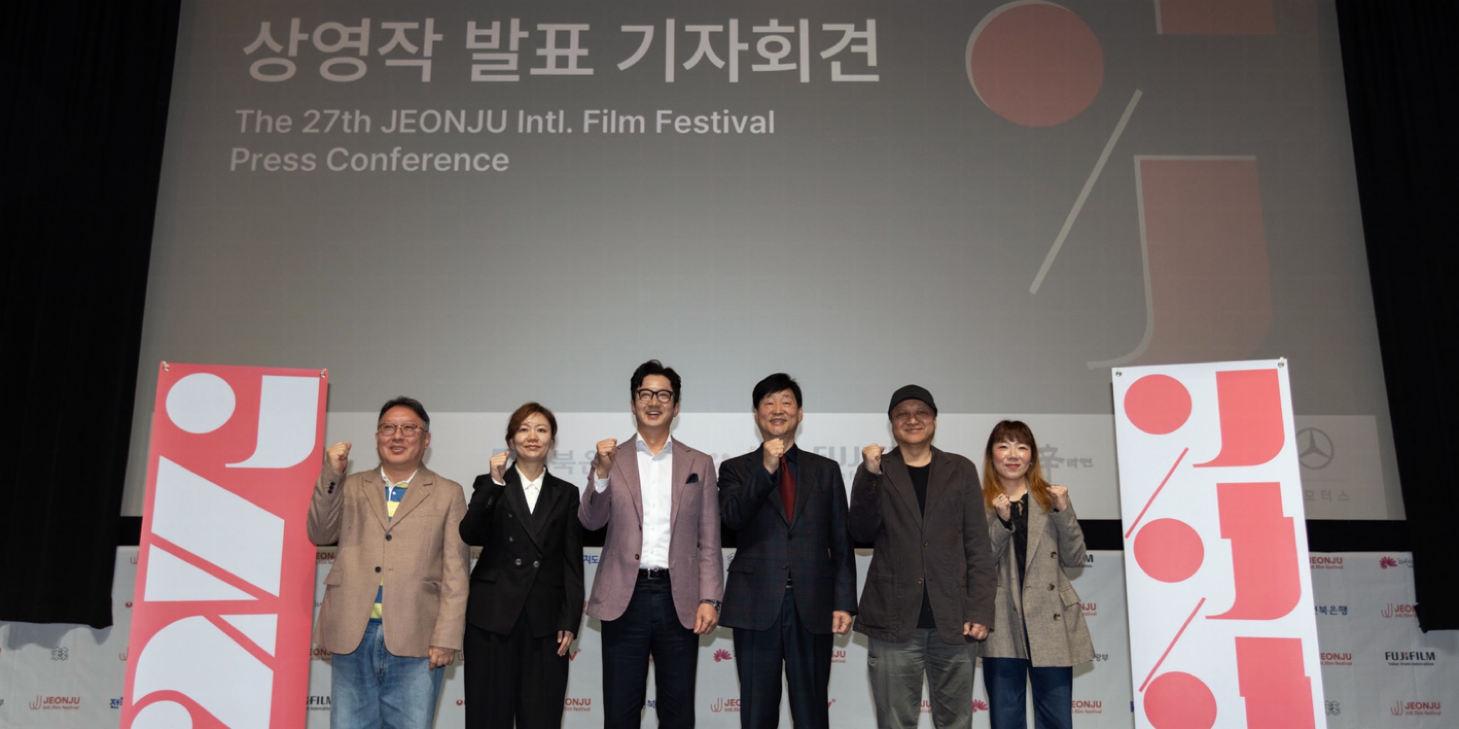 لقطة واسعة من الأمام تظهر ستة أشخاص واقفين على خشبة مسرح بملابس رسمية وبذلات، يرفعون قبضات أيديهم اليسرى عالياً في لفتة تأييد جماعية، أمام شاشة ضخمة بيضاء تعرض شعار مهرجان JEONJU International Film السينمائي الدولي الـ 27 ونصاً باللغتين الكورية والإنجليزية.